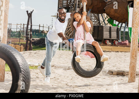 Happy african american father poussant fille sur balançoire pneu à amusement park Banque D'Images