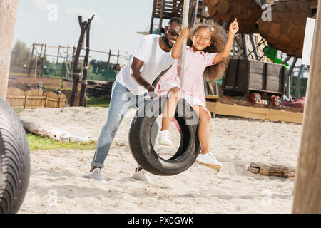 African American father poussant fille sur balançoire pneu à amusement park Banque D'Images
