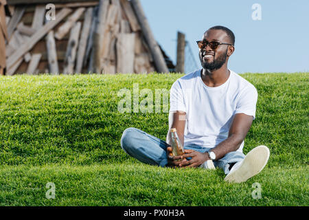 Handsome smiling african american man in sunglasses assis avec soda en bouteille de verre sur la colline verte Banque D'Images