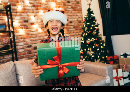 Petite fille surpris montrant sa grande boîte-cadeau en vert et rouge ruban. belle enfant tenant présent montrant de grandes dents sourire. famille à Noël Banque D'Images