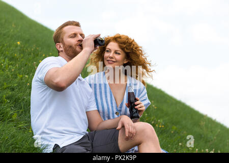 L'homme roux aux yeux clos boisson bouteille de près de smiling girlfriend on grassy hill Banque D'Images