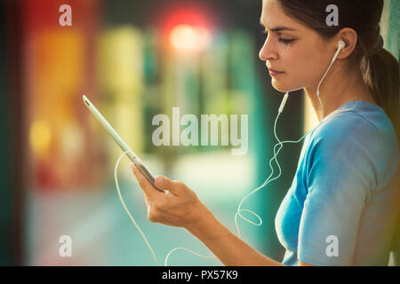 Belle jeune femme est holding a tablet pc, wearing earphones comité permanent par un mur de ciment Banque D'Images