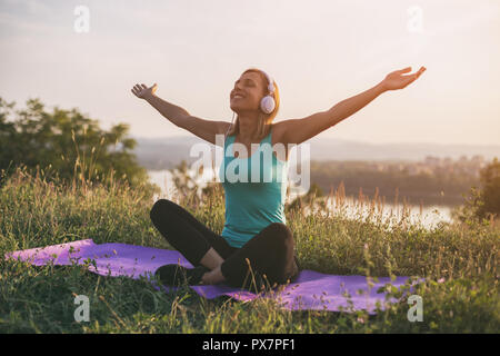 Belle femme sportive avec un casque d'écoute et d'élever la musique bénéficie d'armes tout en restant assis sur tapis d'exercice avec un paysage urbain et sa rivière ces delicous. Banque D'Images