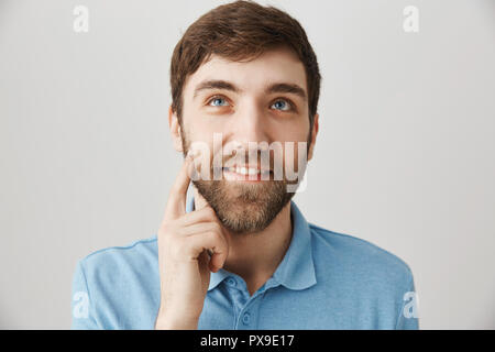 L'Heureux guy rêver ou penser à quelque chose de positif, à l'éraflure et souriant, tout en se tenant la joue contre l'arrière-plan gris. L'homme dans l'amour se rappelle comment petite amie l'embrassa Banque D'Images