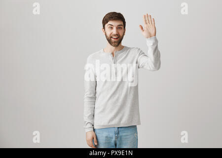 Bonjour mon ami. Tourné à l'intérieur d'ordinaires sympa male office worker waving at camera avec large sourire, voyant un collègue et voulant dire salut, debout sur fond gris Banque D'Images