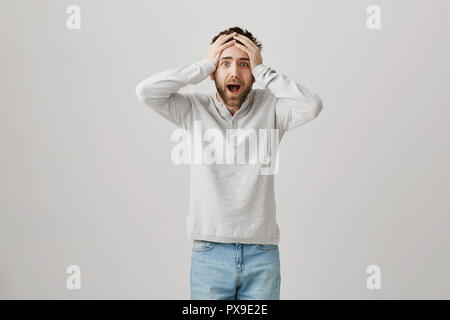 Terrifié misérable barbu tenant les mains sur les cheveux tout en criant et l'expression de l'anxiété et la nervosité, debout sur fond gris. L'homme tremble en voyant son chat marcher sur la rampe de balcon Banque D'Images