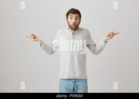 On essaie de regarder dehors. Portrait de confus et remis en question l'homme avec les mains et propagation barbe dirigées dans les deux côtés avec l'expression troublée comme s'il ne sait rien, posant sur fond gris Banque D'Images