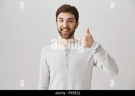 L'émotion positive employé avec sourire brillant showing Thumbs up et d'être dans la bonne humeur tout en se tenant à l'arrière-plan gris. Mate pense que c'est excellente idée de jouer à des jeux Banque D'Images