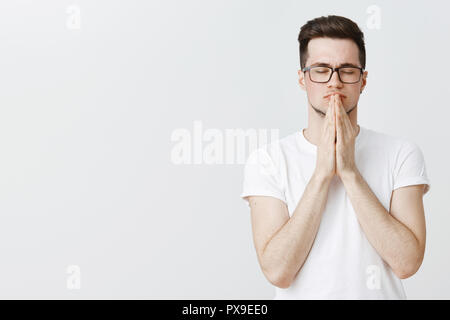 Jusqu'à la taille de balle porté décontracté jeune homme à lunettes avec les yeux fermés essayant calmer holding hands in prier sur lèvres méditation, trouver la paix et la relaxation la pratique du yoga, se libérer du stress Banque D'Images