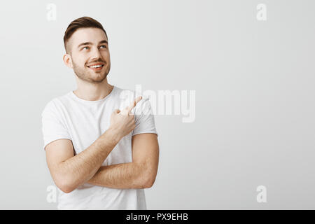 Portrait de l'homme macho charmant et élégant en soie avec hairstyle pointant, contemplant le coin supérieur droit avec plaisir et satisfaction smiling profiter de regarder cool perfomance sur mur gris Banque D'Images