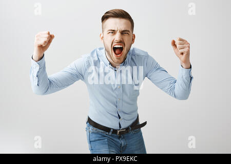 Emotive heureux concernés européenne homme boss en chemise bleue et formelle de flexion jeans vers la caméra en train de crier et de sensibilisation pour encourager l'équipe de football de poings faire bet être en colère sur mur gris Banque D'Images