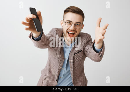 Taille plan sur excité et ambitieux emotive beau mec avec des lunettes et une veste en soie mains tirant vers la caméra profiter de la musique sans fil dans les écouteurs holding smartphone Banque D'Images