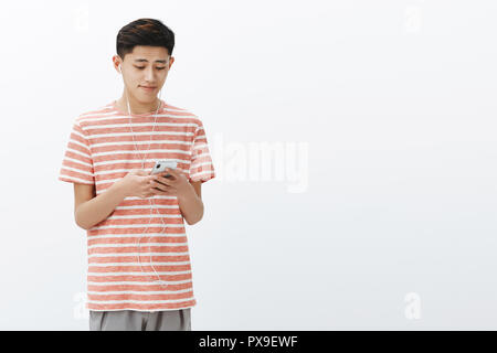 Cute young asian boy in striped t-shirt holding smartphone wearing earphones à mignon et touché à l'écran du téléphone cellulaire, à l'écoute de chanson hearwarming avec coeur de fusion de musique sur mur blanc Banque D'Images