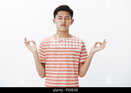 Jusqu'à la taille de balle centrée calme belle asian male student taking control de pensées, standing in lotus pose avec geste zen, méditant la restauration de l'énergie avec le yoga, ce qui pose plus pacifiques mur blanc Banque D'Images