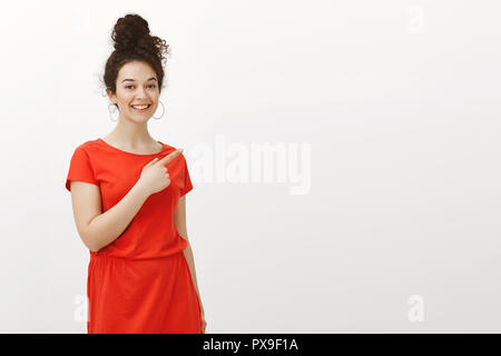Portrait de plaisir jolie femme aux cheveux bouclés en belle robe, pointant vers le coin supérieur droit en souriant largement, l'expression de l'attitude sympathique et heureux, montrant sur fond gris Banque D'Images