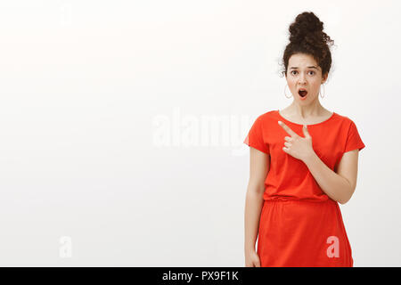 Studio shot of déçu choqué petite amie avec des cheveux bouclés peignées à bun, wearing red dress, passant d'un choc de la mâchoire en pointant le coin en haut à gauche, d'être étonné et stupéfait sur mur gris Banque D'Images