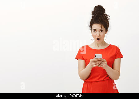 Wtf qui a écrit ce message. Portrait de choqué déplut féminine charmante jeune fille en robe rouge, passant de la mâchoire surprise en holding smartphone et à regarder l'appareil photo, d'être surpris avec news Banque D'Images