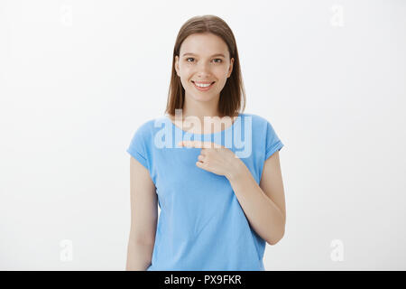 Intrigué joyeux jolie femme avec les cheveux clairs dans des vêtements décontractés, pointant vers la gauche avec l'index et souriant de plaisir curieux expression, discuter de personne à partie, lui demandant s'il datant quelqu'un Banque D'Images