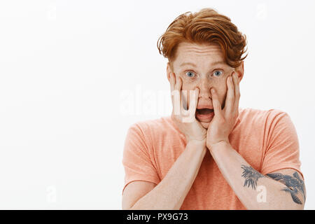 Portrait of cute troublée et intense avec des hommes, les tatouages et les taches de rousseur, cheveux gingembre holding paumes sur les joues et la mâchoire tomber de peur, se sentir nerveux, peur d'être seul à la maison Banque D'Images