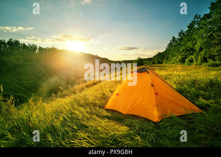 Tourisme Orange tente sur une colline au-dessus du ravin. tente se trouve dans l'épaisse herbe verte au coucher du soleil. Paysage d'été. Le concept de la liberté de déplacement et de pr Banque D'Images