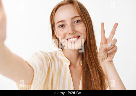 Jusqu'à la taille de balle heureuse et sans soucis mignon jeune rousse femelle avec rousseur en robe jaune tirant part comme si tenir l'appareil photo en souriant et selfies montrant la paix ou la victoire Banque D'Images