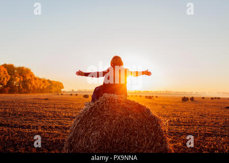 Happy middle-aged woman sitting on haystack en automne et se sentir libre avec les bras ouverts. Se détendre et admirer la nature au coucher du soleil Banque D'Images