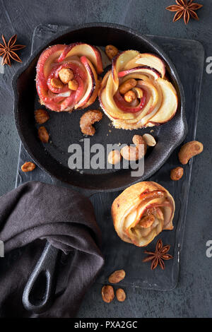 Trois des feuilletés en forme de rose avec des tranches de pomme au four dans une poêle en fonte. Haut lay sur fond sombre avec des draps et serviettes noix caramélisées Banque D'Images