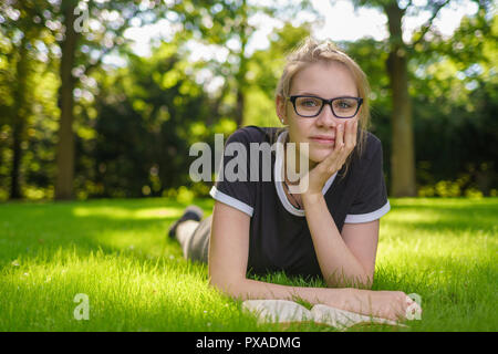 Une jeune femme avec des lunettes se trouve sur l'herbe dans le parc et l'air heureux à l'appareil photo Banque D'Images