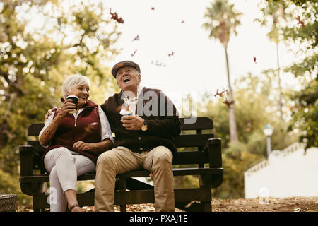 Smiling senior homme et femme assis sur banc de parc avec une tasse de café et de rire. Couple de personnes âgées bénéficiant de pique-nique au parc. Banque D'Images