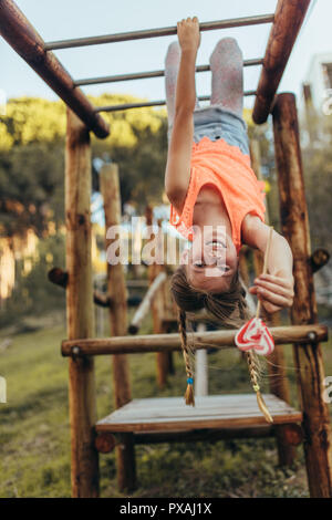 Girl hanging tête en bas dans un parc holding a lollipop candy. Fille jouant dans un parc, tête en bas sur une échelle horizontale bénéficiant d'une Banque D'Images