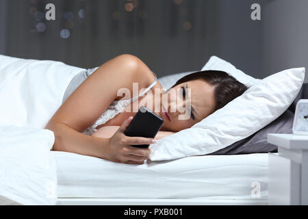 Femme en colère contrôle de contenu smart phone sur le lit dans la nuit à la maison Banque D'Images