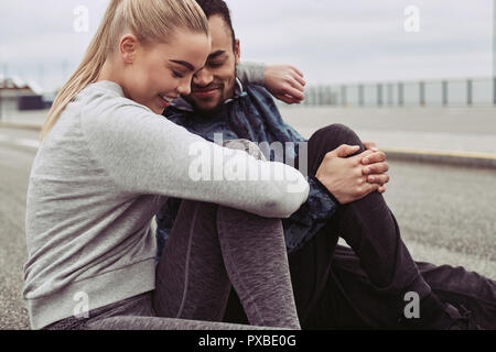 Diverses jeune couple assis sur un chemin ensemble et souriant tout en faisant une pause à partir d'une exécution sur un jour nuageux Banque D'Images