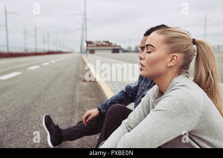 Jeune couple divers dans les vêtements de sport assis sur le bord d'une route ensemble en faisant une pause à partir d'une exécution sur un jour nuageux Banque D'Images