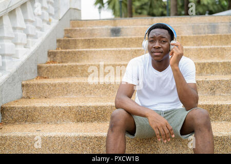 Beau jeune homme africain assis sur des escaliers tout en écoutant de la musique avec des écouteurs Banque D'Images