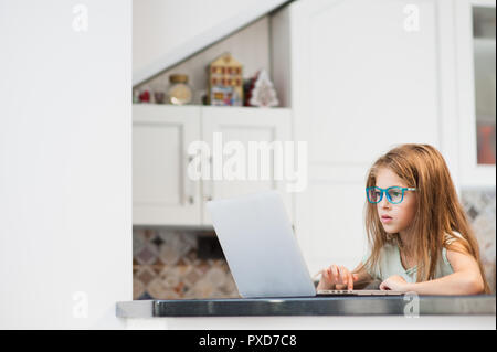 Belle petite fille dans les verres dans la cuisine en appuyant sur le clavier sur un ordinateur portable sans fil Banque D'Images