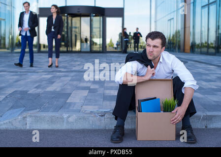 Tiré business man sitting frustré et contrarié dans la rue près de l'immeuble de bureaux de fort de ses affaires. Il a perdu travail Banque D'Images