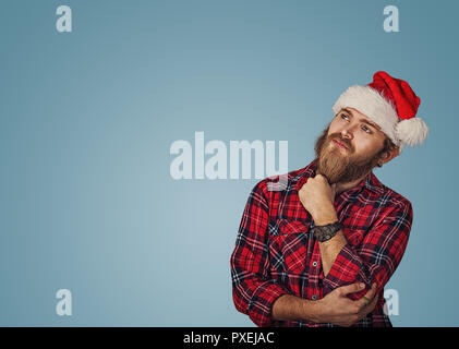 Beau jeune homme barbu hippie dans Christmas hat looking up posing in studio pense à idées cadeaux isolé sur fond bleu mur studio avec co Banque D'Images