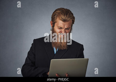 L'homme d'affaires malheureux homme barbu holding laptop computer fronçant isolé sur fond gris gris mur studio. Les droits de l'expression négative face emotio Banque D'Images