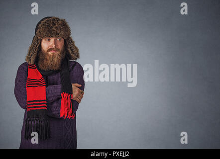 J'ai froid. Homme barbu à chapeau de fourrure russes moelleuses, jeune homme faire de geste à froid sur le côté pour copie espace isolé sur mur gris gris studio B Banque D'Images