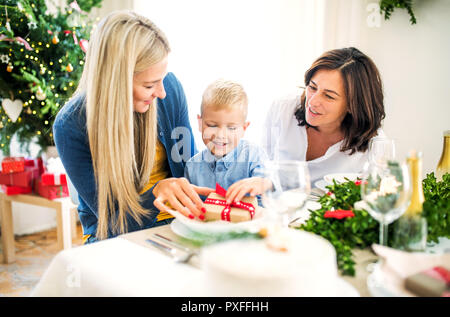 Un petit garçon avec le présent et mère et grand-mère assis à une table à la maison au moment de Noël. Banque D'Images