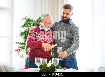 Un père et fils adultes stabding à une table, verser le vin dans un verre au moment de Noël. Banque D'Images