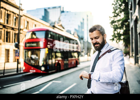 Hipster businessman standing on the Street à Londres, contrôle du temps. Un douuble bus rouge à deux étages à l'arrière-plan. Banque D'Images