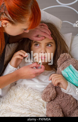 Une petite fille avec son ours en peluche préféré sur lequel elle portait un bandage de gaze est malade au lit et sa mère ses mesures avec une température elec Banque D'Images