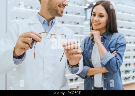 Vue partielle de l'oculiste mâle heureux d'aider les femmes à choisir paire de lunettes en magasin ophtalmique Banque D'Images