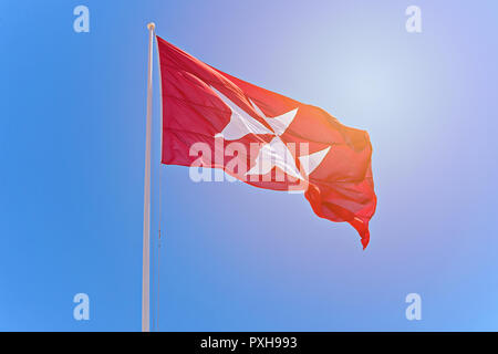 Drapeau rouge avec croix noire. Symbole de l'Ordre des Chevaliers de Malte est la croix à huit pointes au lever du soleil sur fond de ciel bleu, l'espace de copie, soufflant dans le vent Banque D'Images