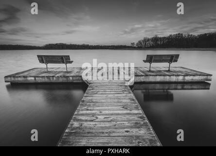 A lonely pier en noir et blanc avec deux bancs assis sur un lac calme. Banque D'Images