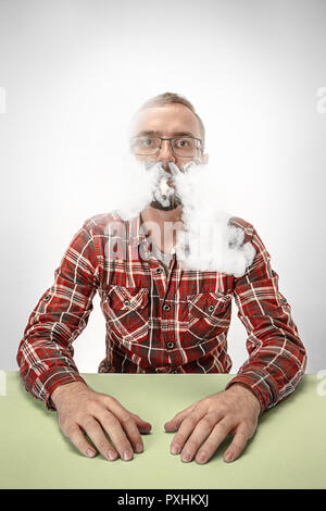 Beau hipster man smoking cigarette et assis à table à la maison ou en studio. L'homme regardant vers le haut et profiter de passer du temps libre. Banque D'Images