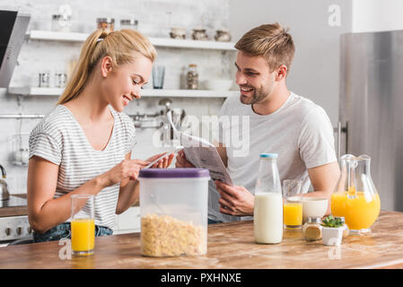 Petite amie en utilisant smartphone et petit ami reading newspaper in kitchen Banque D'Images