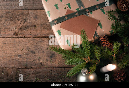 Des brindilles de sapin vert, des pommes de pin et des boules de Noël, près d'un cadeau enveloppé dans du papier brun classique avec des arbres, et une bougie allumée, sur une table en bois vintage. Banque D'Images