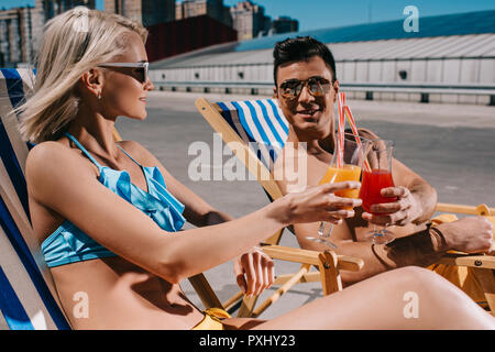 Jeune couple clinking glasses de cocktails tout en étant assis sur des chaises longues sur le stationnement Banque D'Images